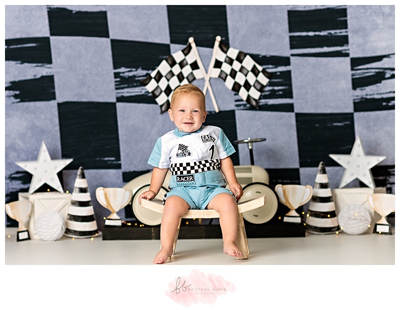 One year old boy sitting on white stool smiling in front of race-themed cake smash set