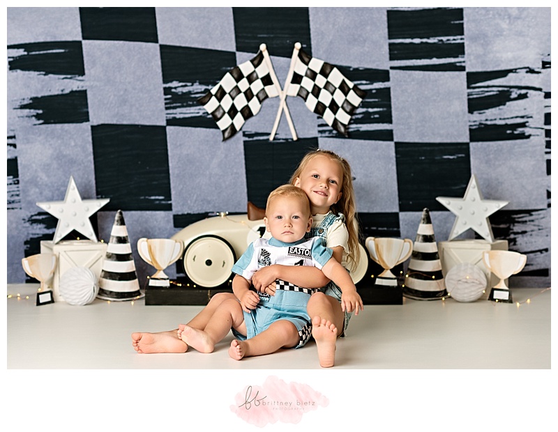 One year old boy sitting with big sister in front of race-themed cake smash set