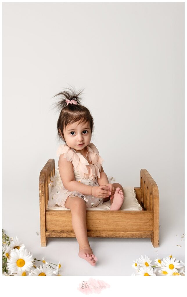  One-year-old baby girl with a bunch of daisies and sitting on a bed during a cake smash photography session in Calgary