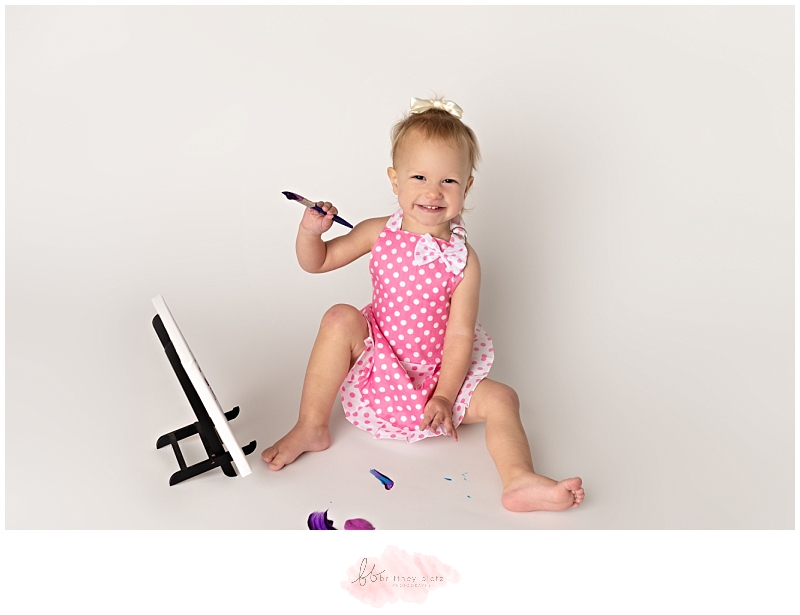 2 year old in pink, polka dot apron smiling while painting on canvas