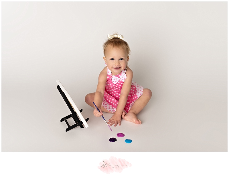 2 year old in pink polka dot apron dipping paint brush into paint